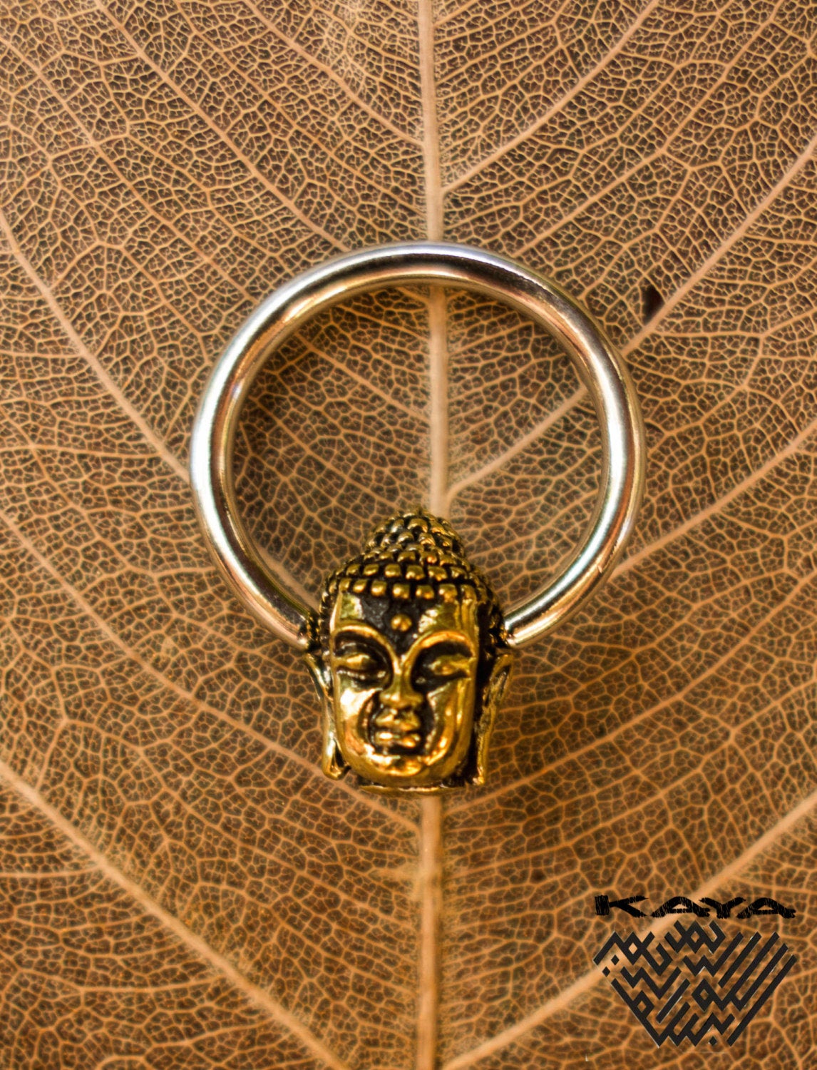 A BUDDHA Silver Ring with a Gold Pendant from Kaya Movement is elegantly placed on a dried leaf with its intricate veins visible. The gold brass Buddha head sits in the center of the silver circular band, exuding an essence of serenity and elegance. The word "kava" appears in the bottom right corner, adorned with a decorative pattern.